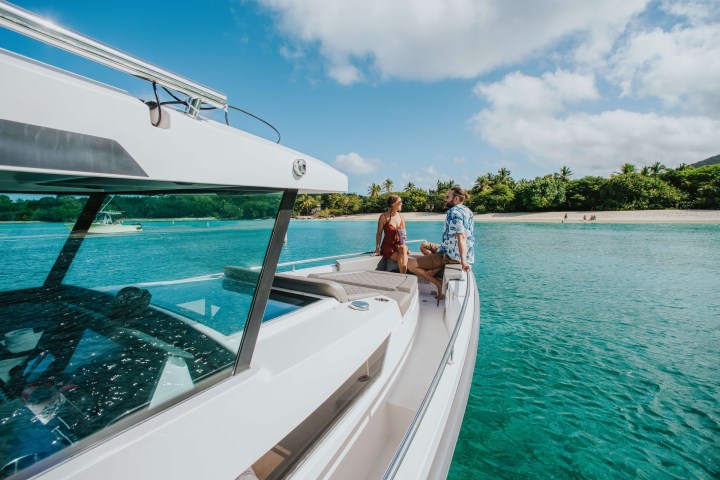 Two people standing on a boat near a tropical beach with clear blue water and palm trees.