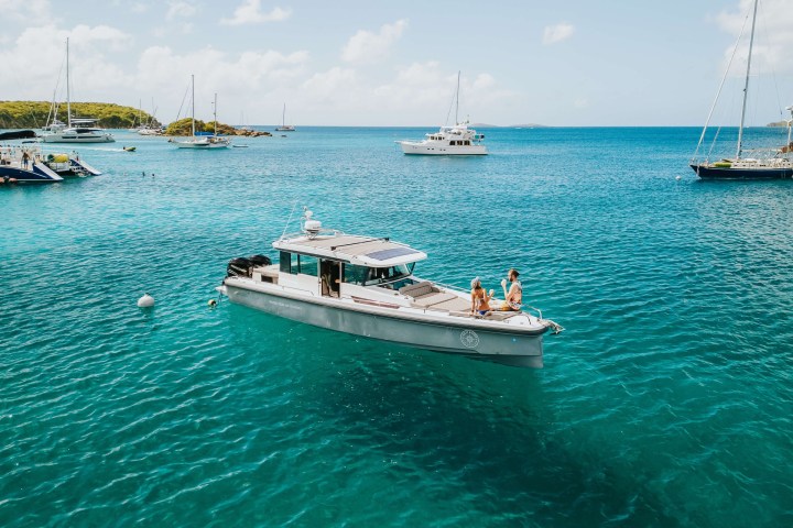 Motorboat with people in a sunny bay, surrounded by sailboats and clear blue water.